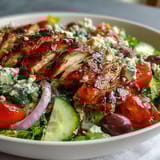 Grilled chicken breast slices over crisp Romaine lettuce, cherry tomatoes, and cucumbers in a classic Greek salad bowl.  