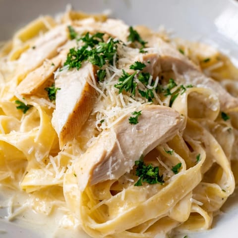 Steaming fettuccine Alfredo pasta coated in a creamy Parmesan sauce, topped with sliced sautéed chicken breast on a white plate.